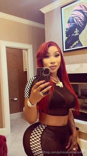 Woman with red hair in sheer black and checkered outfit, taking mirror selfie in indoor setting.