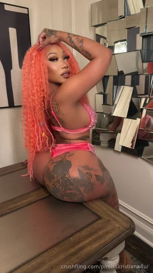 Woman with bright orange hair and tattoos wears pink lingerie, posing on a wooden bench indoors.