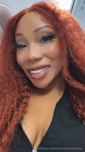 Woman with curly red hair wearing a black zip-up top, smiling in a close-up selfie.
