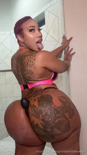 Woman with tattoos in pink lingerie posing in a bathroom, showing back and buttocks.