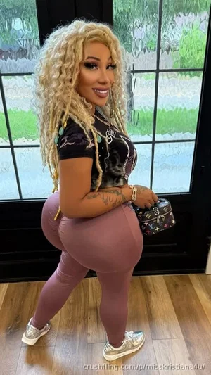 Woman with curly blonde hair in tight pink leggings and black top, holding a small bag indoors near