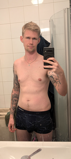 Man with tattoos taking mirror selfie in bathroom wearing boxers.