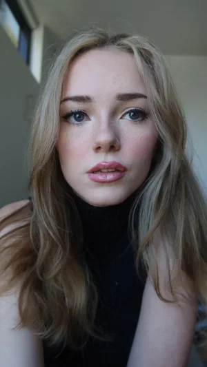 Blonde woman in black top, indoor close-up selfie, soft lighting.