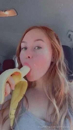 Woman with long blonde hair eating a banana in a car, wearing a gray top.
