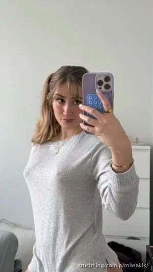 Woman in light gray long-sleeve top taking mirror selfie indoors.