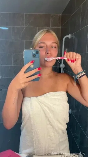 Blonde woman in towel brushing teeth in bathroom mirror selfie.
