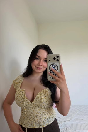 Woman with long dark hair in floral top and dark pants, taking mirror selfie in bedroom.
