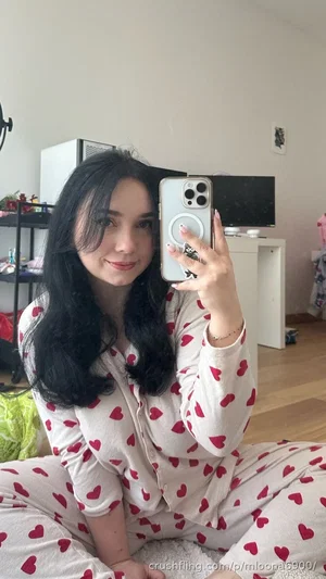 Woman with long dark hair in heart-patterned pajamas taking mirror selfie in bedroom.