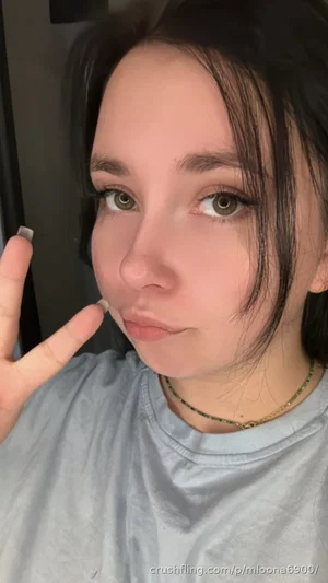Woman with dark hair and green eyes wearing a gray t-shirt, making a hand gesture indoors.