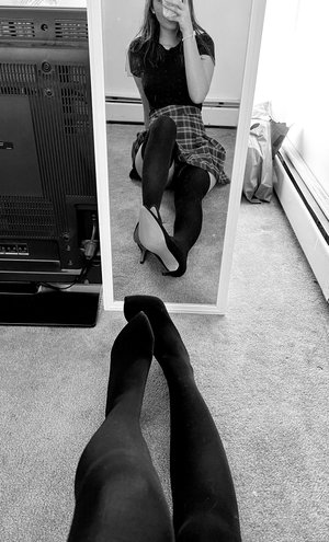 Woman in black top, plaid skirt, stockings, heels, mirror selfie.