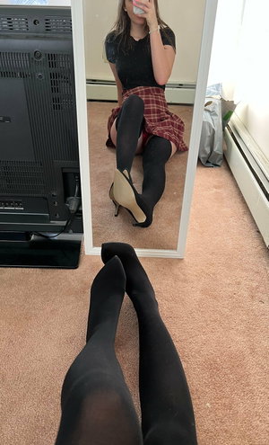 Woman in black top, plaid skirt, stockings, heels, mirror selfie.