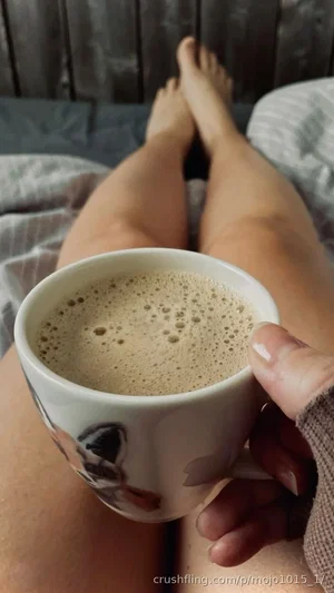 Woman holding a mug of coffee, legs visible on a bed, indoor setting.