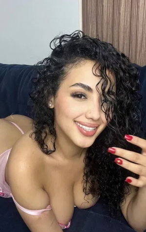 Woman with curly black hair in pink lingerie lying on dark couch, smiling at camera.
