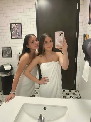 Two women in a bathroom, one wrapped in a white towel, taking a mirror selfie.