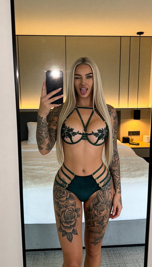 Woman with long blonde hair and tattoos wears green lingerie, takes mirror selfie in bedroom.