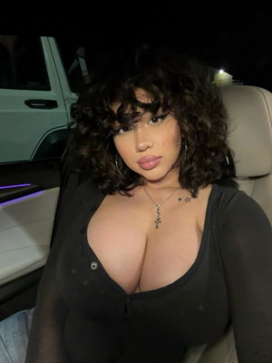 Woman with curly hair in black sheer top sitting in car at night.