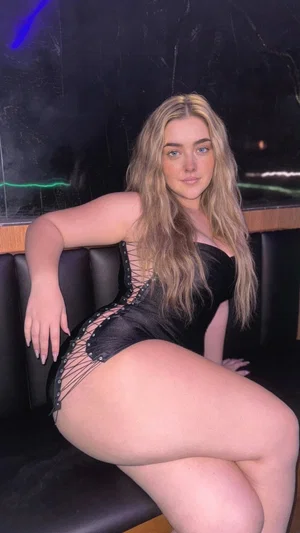 Woman with long blonde hair in black lace-up bodysuit posing on dark couch indoors.
