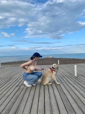 Topless woman in jeans and cap squats with dog on seaside pier.