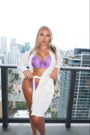 Woman in purple lingerie and white robe posing on balcony with city view.