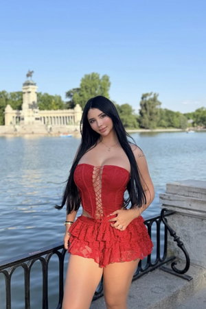 Woman in red lace corset and skirt posing by water near monument.