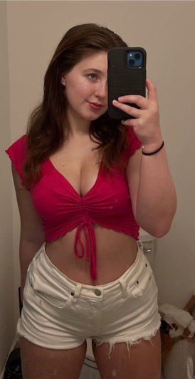 Woman in pink crop top and white shorts taking mirror selfie indoors.