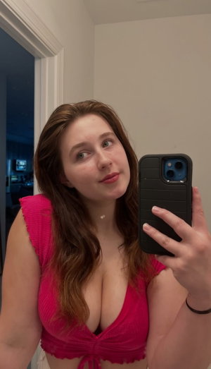 Woman in pink top taking mirror selfie indoors, long brown hair, smiling.