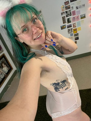 Woman with teal hair and glasses wears white lace lingerie in bedroom selfie.