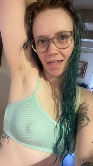 Woman with green hair and glasses wears light green bra, showing tattoos and armpit hair in indoor