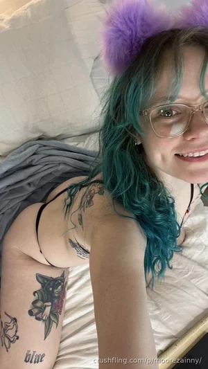Woman with teal hair and glasses, wearing black bra, lying on bed with tattoos visible.