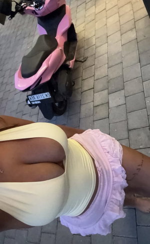 Woman in yellow top and pink ruffled shorts standing near pink scooter on brick pavement.