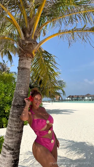 Woman in pink swimsuit posing by palm tree on sandy beach.