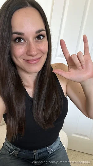 Woman with long brown hair in black tank top and jeans, smiling and making hand gesture indoors.
