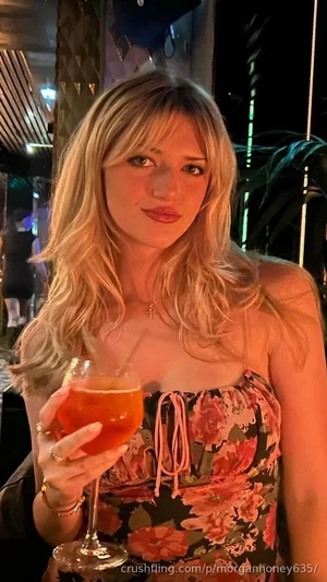 Blonde woman in floral top holding a drink at a dimly lit indoor venue.