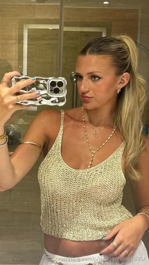 Woman with blonde ponytail in beige knit top and white skirt, taking mirror selfie in bathroom.