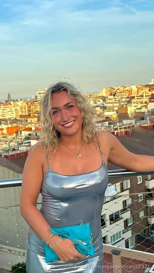 Woman with curly blonde hair in silver dress holding blue clutch on balcony overlooking city.