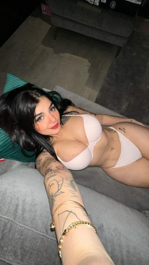 Woman with dark hair and tattoos wears white lingerie, lying on gray couch indoors.