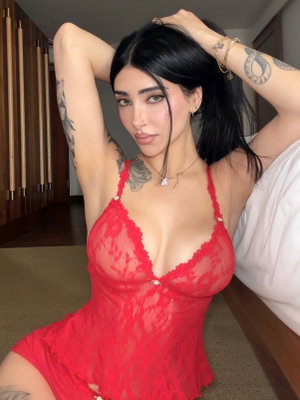 Woman in red lace lingerie with tattoos, posing indoors near bed.