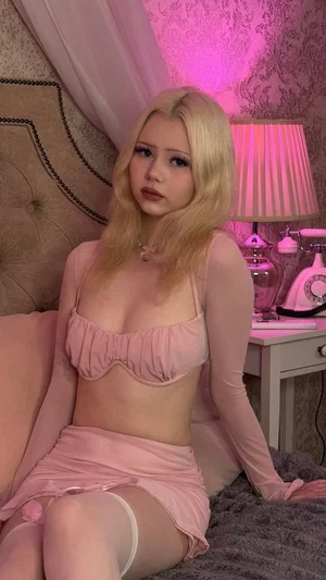 Blonde woman in pink lingerie and sheer sleeves sitting on bed in pink-lit bedroom.