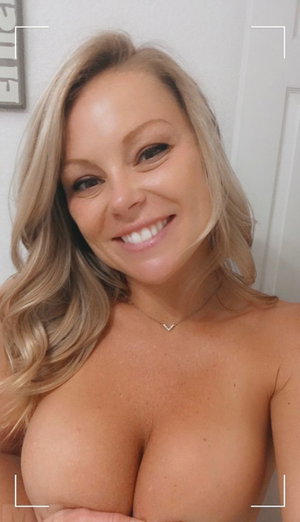 Blonde woman smiling in a topless selfie indoors.