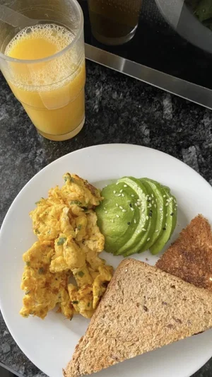 Plate with scrambled eggs, avocado slices, toast, and a glass of orange juice on a dark countertop.
