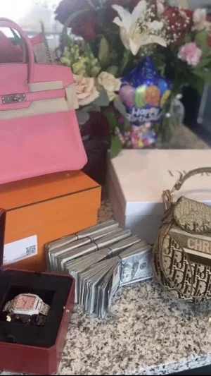 Luxury items on a countertop: pink handbag, cash, watch, flowers, and a Dior bag.