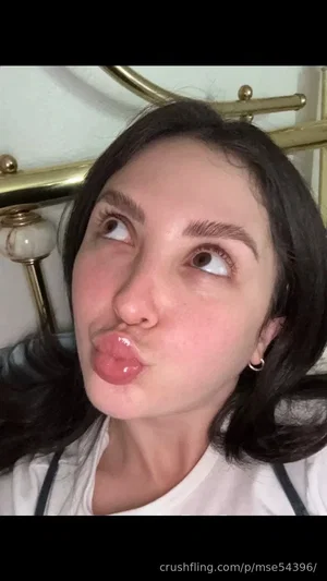 Woman with dark hair making a playful face, wearing a white top with black straps, indoors near a