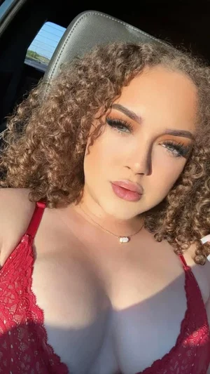 Woman with curly hair in red lace top sitting in car.
