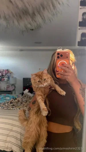 Woman with tattoos holding a ginger cat in a bedroom, wearing a brown crop top and matching pants.