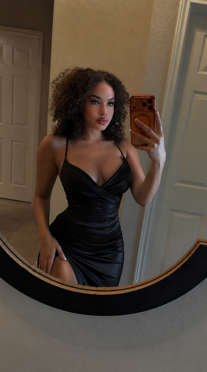 Woman with curly hair in black dress taking mirror selfie indoors.