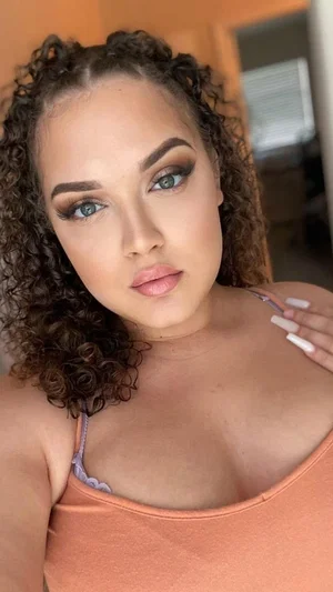 Woman with curly hair and makeup wearing an orange tank top indoors.