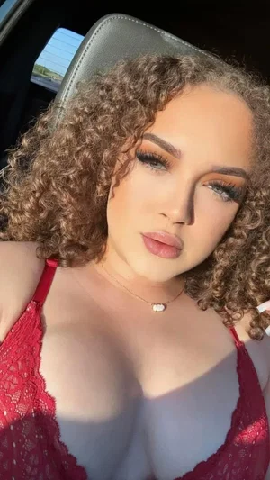 Woman with curly hair in red lace top sitting in car, wearing necklace.
