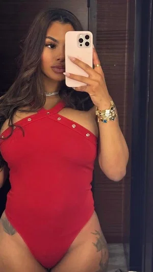 Woman in red one-piece swimsuit with cutouts, tattoos on thighs, taking mirror selfie indoors.