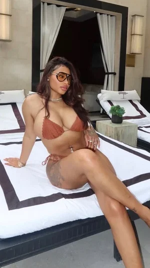 Woman in brown bikini posing on bed in bedroom, wearing glasses and jewelry.