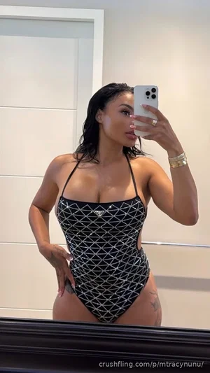 Woman in black patterned one-piece swimsuit taking mirror selfie indoors.
